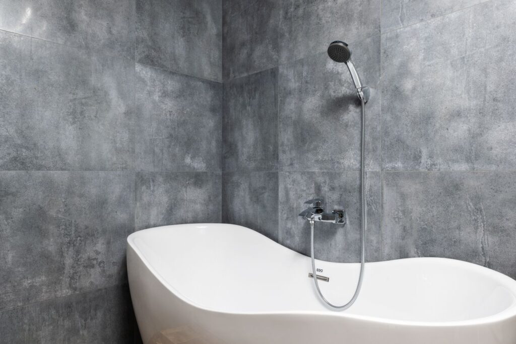 Interior of light modern bathroom with white bath under shower on gray tile wall